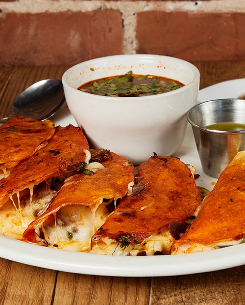 Four birria quesa tacos with melted cheese and a white bowl of consome, on a wooden table in front of an exposed red brick wall