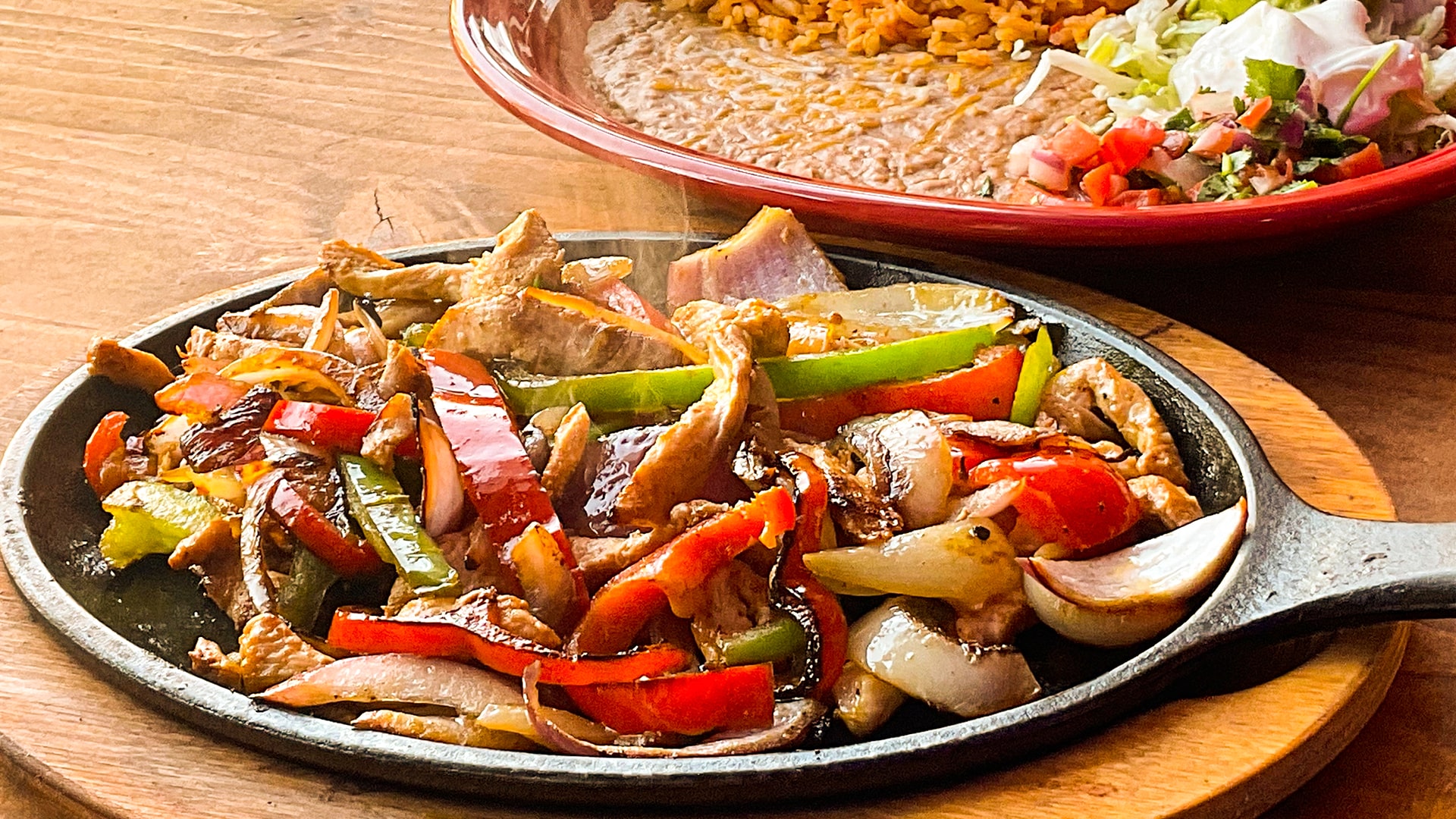 Chicken fajitas with red and green peppers and onions steaming on a black cast iron comal, a red plate of rice, beans and pico behind